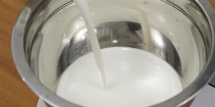 Close-up shot of liquid white milk being poured in a steady stream into a clean, reflective stainless steel mixing bowl. The slow movement captures the smooth texture of the dairy liquid pooling at th