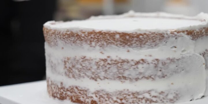 A detailed, close-up shot capturing a professional confectioner smoothing creamy white buttercream frosting onto the sides of a fresh three-layer sponge cake using a metal palette knife. Perfect for b