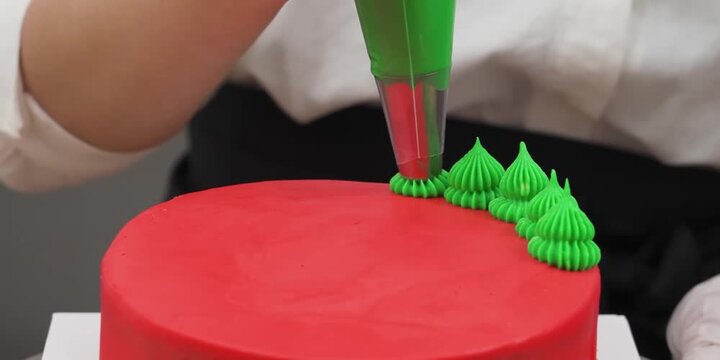 Close-up shot of a pastry chef using a piping bag to create small, decorative green buttercream trees on the top edge of a vibrant red frosted cake. Perfect for holiday baking, Christmas dessert prepa