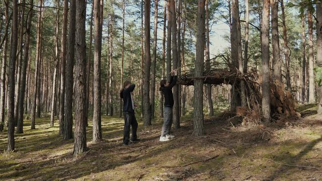 Two teenagers build a house out of branches. The long-haired brothers stand near a wooden lean-to in a sunlit clearing in a temperate forest. Sunbeams filter through the leaves