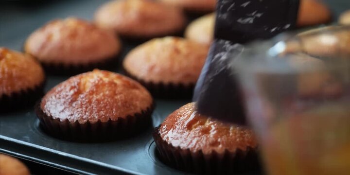 A close-up shot of a baker using a pastry brush to apply a glossy syrup glaze onto warm, freshly baked golden muffins in a muffin tin.