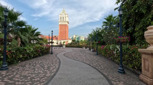 Wide shot of a beautiful alley in a Vietnamese park featuring a mosaic-paved path, manicured flower bushes, palm trees, and a large clock tower in the background.