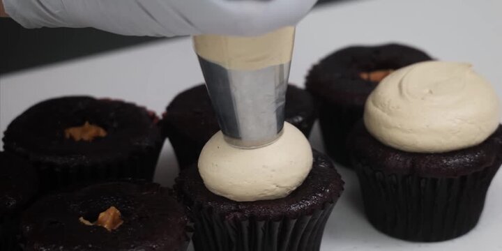 A gloved baker uses a pastry bag with a large round tip to pipe smooth, creamy frosting onto freshly baked chocolate cupcakes. The cupcakes feature a distinct caramel or dulce de leche core filling vi