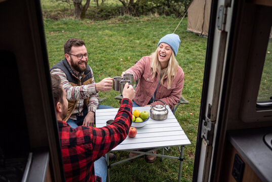 Group of Cheerful Friends Toasting with Metal Mugs at Camping Table Outside Campervan