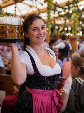L&auml;chelnde Bedienung im Dirndl serviert Getr&auml;nke am Abend auf einem deutschen Volksfest, w&auml;hrend ein beleuchtetes Riesenrad und Fahrgesch&auml;fte f&uuml;r stimmungsvolle Kirmesatmosph&auml;re sorgen.