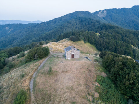 Aerial view of a stone chapel on a grassy hilltop in the Parco Nazionale dell'Appennino Tosco-Emiliano, surrounded by dense forests and mountain slopes in Massa-Carrara, Italy.