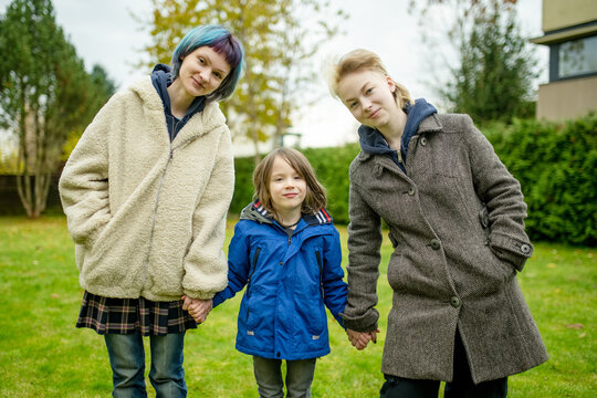 Three siblings pose together outdoors on a chilly autumn day, with a smiling boy hugging his older sister. Natural family portrait in a garden setting.