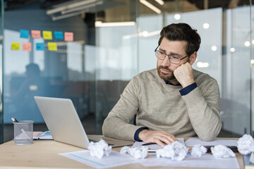 Fototapeta premium Stressed man experiencing mental exhaustion and professional burnout, resting his head on his hand while sitting at a laptop-filled desk with crumpled papers in a modern office environment