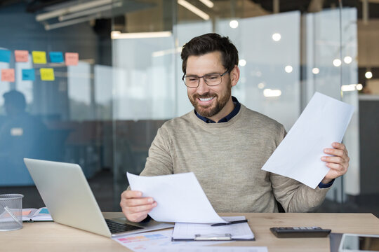 Happy businessman reviewing financial paperwork, smiling while managing reports and using a laptop at his modern office desk, feeling successful and organized