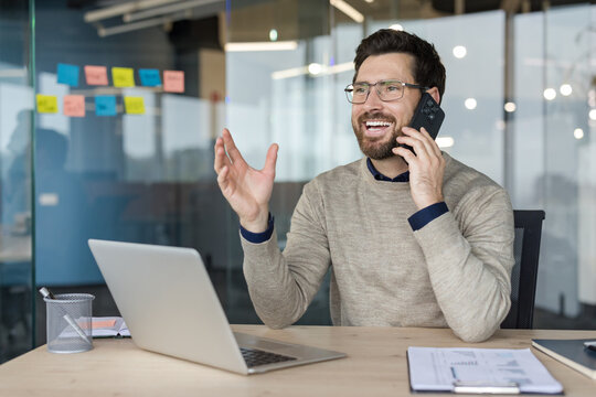 Smiling man communicating on a mobile phone while working on a laptop at an office desk, discussing business strategies and planning for future success