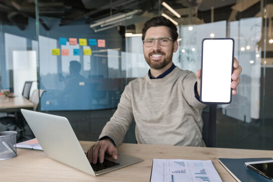 Young businessman in modern office smiling as he holds a smartphone with blank screen toward camera for app mockup, working at laptop surrounded by documents and charts