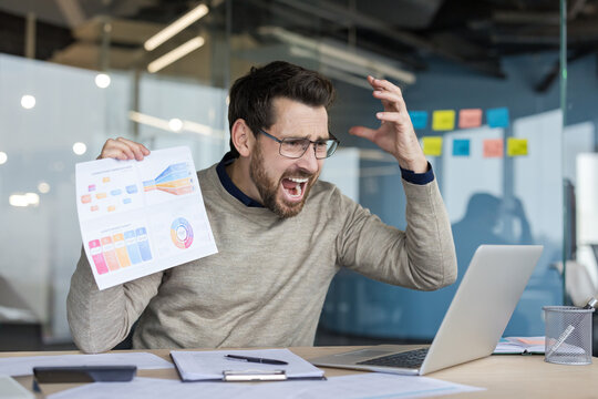 Stressed businessman reacting with frustration and anger while looking at a laptop screen and holding a financial data report, experiencing failure and burnout in the modern office environment