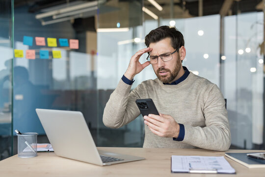 Stressed businessman sitting at an office desk, holding a smartphone, and reacting with a worried expression while receiving bad news or experiencing a problem