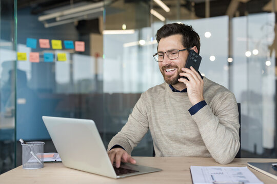 Young man smiling and communicating on a mobile phone while typing on a laptop, performing multiple tasks and staying connected in a modern office environment
