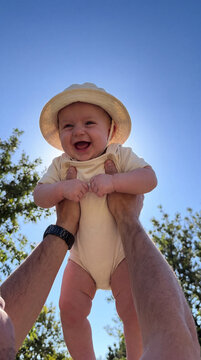 Joyful Baby Wearing Sun Hat Lifted High in the Air by Parent Outdoors on Sunny Day