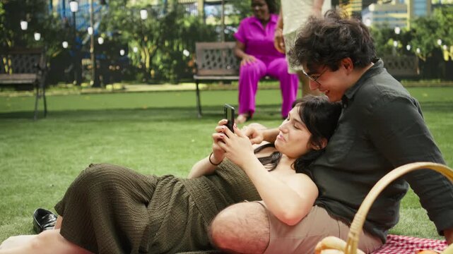 Laughing young woman leans on man and uses a smartphone while sharing amusing reels on a picnic blanket in urban park, as boyfriend and girlfriend enjoy a joyful funny moment during an outdoor date.
