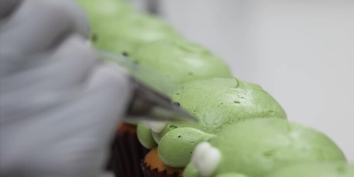 A close-up, high-definition shot showcases a professional baker wearing protective white gloves as they meticulously apply finishing details to a caterpillar-shaped pull-apart cupcake cake. Using a pi