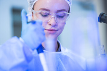 A female microbiologist in a white coat, protective eyewear and a sterile hairnet watching pipette...