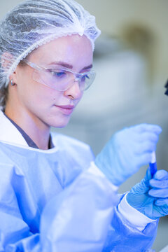 A woman researcher in white laboratory clothing, safety glasses and blue gloves rolling the blood tube between gloved fingers inside a university biotechnology laboratory.
