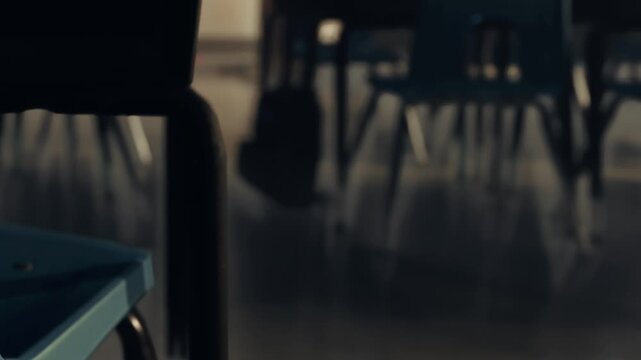 A classroom with several empty chairs and tables. Sunlight enters through windows, casting shadows on the floor. A backpack is positioned in the background.