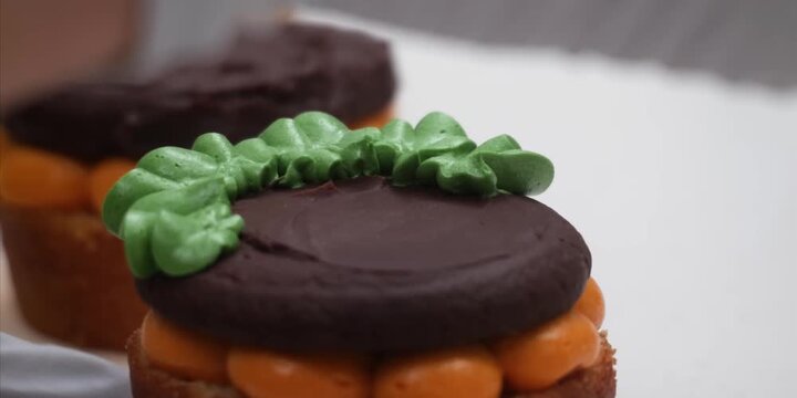 A detailed macro shot capturing the precise culinary artistry of a pastry chef adding a vibrant green buttercream leaf onto a festive, pumpkin-shaped pastry. The gourmet dessert features layers of bri