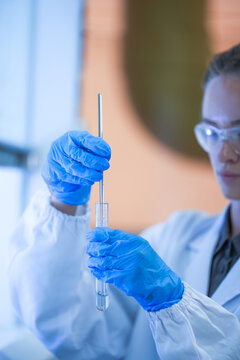 A young female lab scientist in white laboratory clothing, safety glasses and blue gloves holding tube and rod at the hood ledge in a modern medical research laboratory.