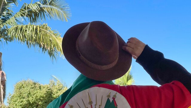 Holding a hat on their head and the flag of Rio Grande do Sul (southern Brazil) across their chest, amidst nature and under a blue sky. Tradition, freedom, and pride in being *ga&uacute;cho*. Farroupilha Wee