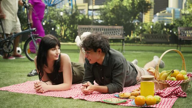 Young couple lies face to face on a picnic blanket and smiles while talking during a relaxed outdoor date with an urban skyline in the background, sharing eye contact and a romantic picnic date.