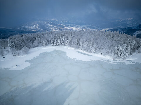 Aerial view of the frozen lake in Parco Nazionale dell'Appennino Tosco-Emiliano, surrounded by snow-covered pine forests under a cloudy winter sky in Sassalbo, Massa-Carrara, Italy.