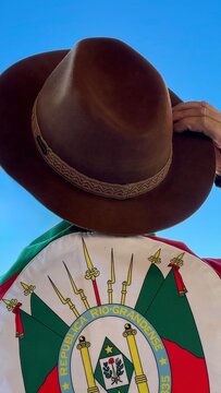 Holding a hat on their head and the flag of Rio Grande do Sul (southern Brazil) across their chest, amidst nature and under a blue sky. Tradition, freedom, and pride in being *ga&uacute;cho*. Farroupilha Wee