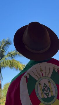 A hat on the head and the flag of Rio Grande do Sul (southern Brazil) across the chest, amidst nature and under a blue sky. Tradition, freedom, and pride in being a *ga&uacute;cho*. Farroupilha Week.