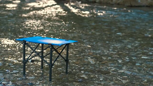 Camping table standing in a river stream. Light blue folding camping table standing in shallow river waters, reflecting bright sunlight across rippling surface, capturing serene wilderness moment