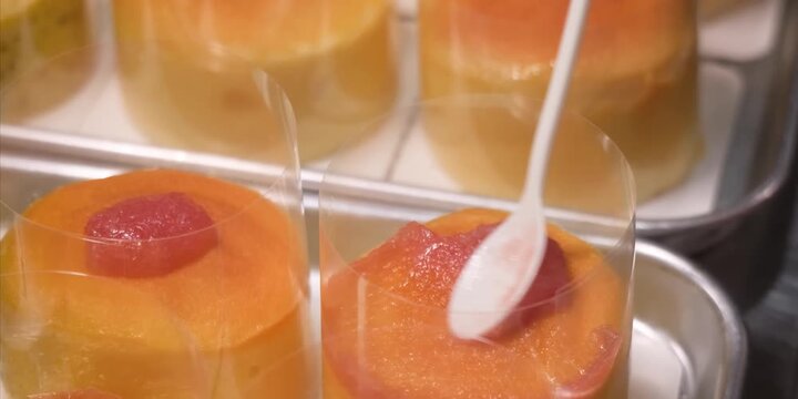 A detailed close-up shot capturing a pastry chef using a spoon to carefully pour a vibrant red fruit glaze or coulis onto individual, multi-layered mango mousse cakes. The mini desserts are neatly arr