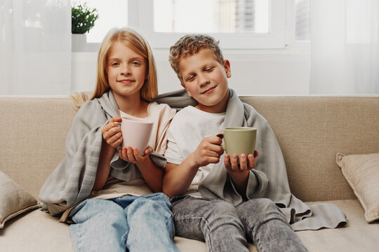 Two children wrapped in blankets sit on a beige sofa holding warm mugs, enjoying a cozy moment in a bright living room with natural light from the window behind them.