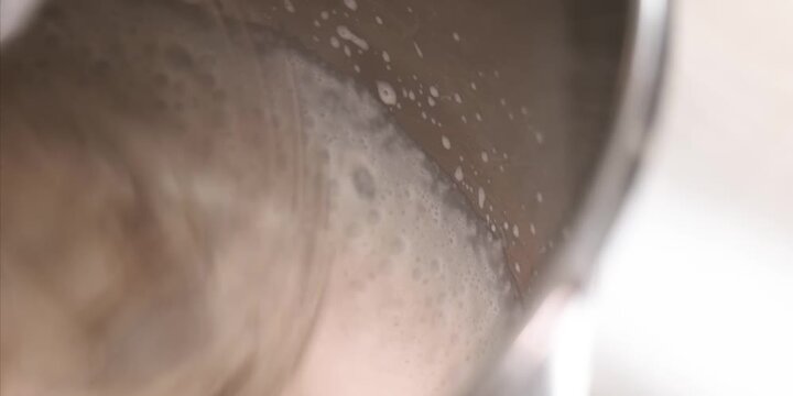 A high-angle, extreme close-up shot captures thick, creamy milk foam swirling smoothly inside a stainless steel steaming pitcher. Tiny, dense bubbles form a soft texture along the edges, making this f