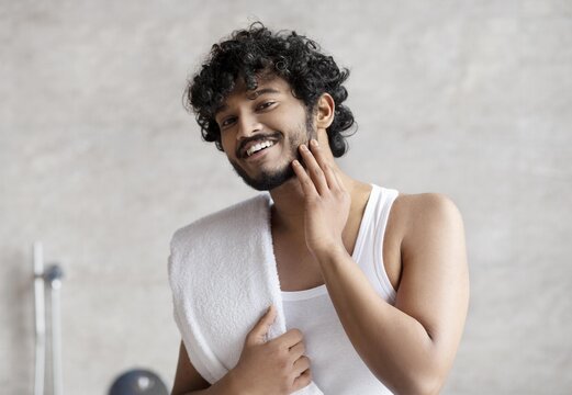 Happy Young Man With Curly Hair Smiling And Touching His Beard After Shaving At Home. Enjoying Skincare Routine In Modern Bathroom Setting. Weight Loss Concept.