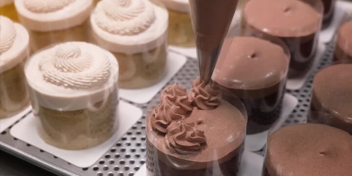 A close-up shot of a professional pastry chef using a piping bag to carefully decorate individual chocolate mousse cakes. The scene features rows of beautifully prepared mini cakes and cupcakes wrappe