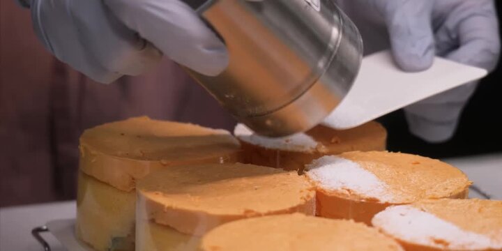 Close-up stock footage of a pastry chef wearing blue protective gloves, using a metal shaker and a white card to carefully dust powdered sugar or icing over a row of small, round orange frosted cakes 