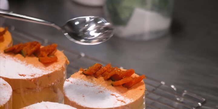 A close-up, high-definition stock video clip showing a pastry chef using a metal spoon to drizzle a clear, glossy sugar syrup over a row of small, round orange cakes. The gourmet desserts feature a sm