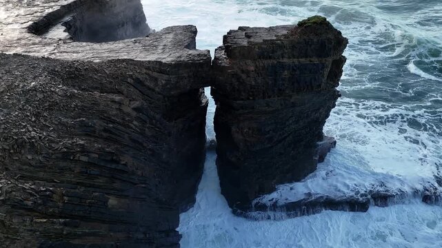 Cinematic aerial view of a lone traveler on a cliff precipice observing rough ocean waves and a majestic sea stack along the Wild Atlantic Way