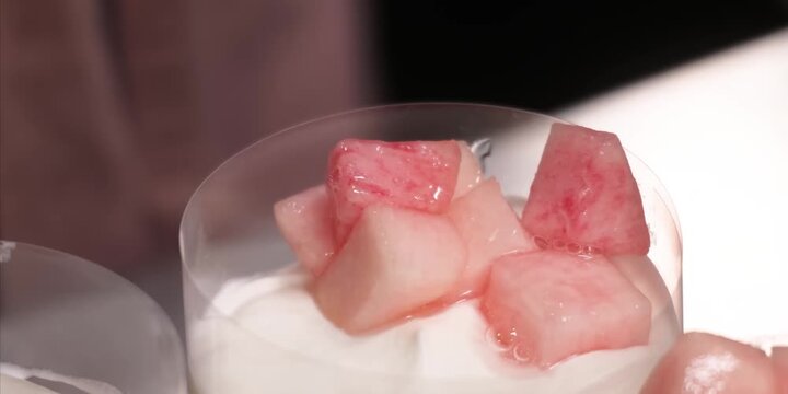 A high-definition, macro tracking shot showcasing succulent, pink-hued diced peaches sitting elegantly atop a rich, velvety white layer of Greek yogurt or cream panna cotta. The soft, cinematic studio
