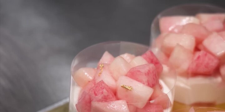 A close-up shot shows a chef wearing a purple nitrile glove using a wooden toothpick to carefully place a delicate piece of edible gold leaf onto a heap of freshly diced, juicy pink and white peaches.