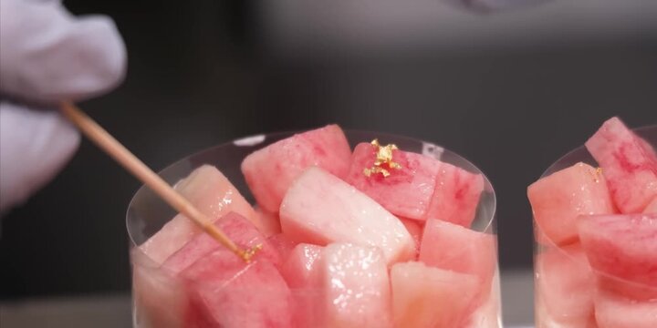 A close-up, shallow depth of field shot of fresh, juicy pink and white diced peach chunks layered inside transparent dessert cups. Small gold leaf flakes delicate adorn the top of the fruit pieces, cr