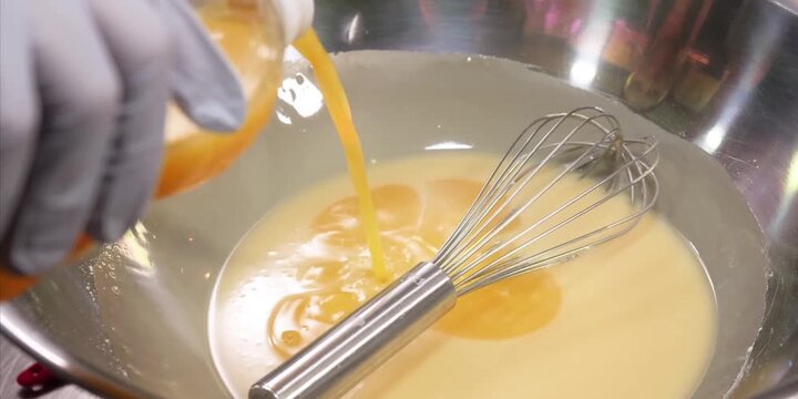 Close-up stock footage of a gloved hand pouring a bright yellow liquid or oil into a light-colored batter inside a large stainless steel mixing bowl. A metal balloon whisk rests in the mixture, ready 