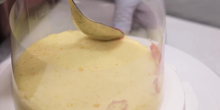 close-up shot of a pastry chef wearing white gloves, using a metal spoon to evenly spread pink fruit cream or filling along the edges of a yellow sponge cake layer. The cake is built inside a clear pl