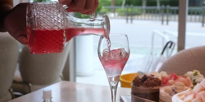 A close-up shot of a hand pouring a vibrant pink beverage or ros&eacute; wine from an elegant, textured crystal carafe into a clear wine glass filled with ice cubes. In the soft-focus background, an array of