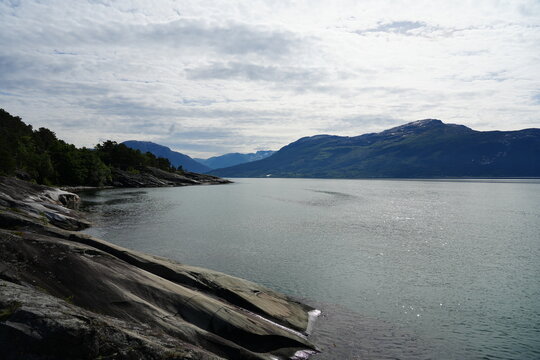 Rocky shoreline overlooking a calm Norwegian fjord and layered mountains beneath a cloudy sky.