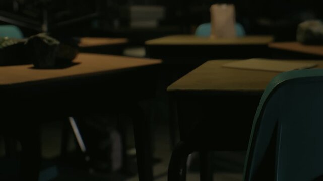 A classroom is dimly lit with empty desks and chairs. A few school supplies lie on one desk. Shadows create a quiet atmosphere. It is evening and no students are present.