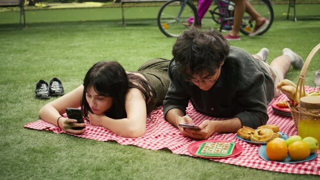 Young couple shows phone addiction and no communication while lying side by side on a picnic blanket and doomscrolling on separate smartphones.