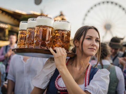 Erfahrene Bedienung im Dirndl tr&auml;gt mehrere gro&szlig;e Bierkr&uuml;ge auf der Schulter &uuml;ber den Festplatz eines deutschen Volksfests &ndash; routinierter Service und traditionelle Festkultur.